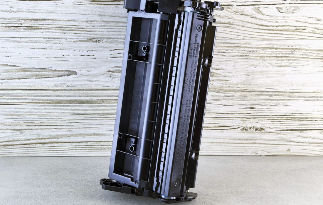 Close-up of a black printer toner cartridge against a rustic wooden background, side view.